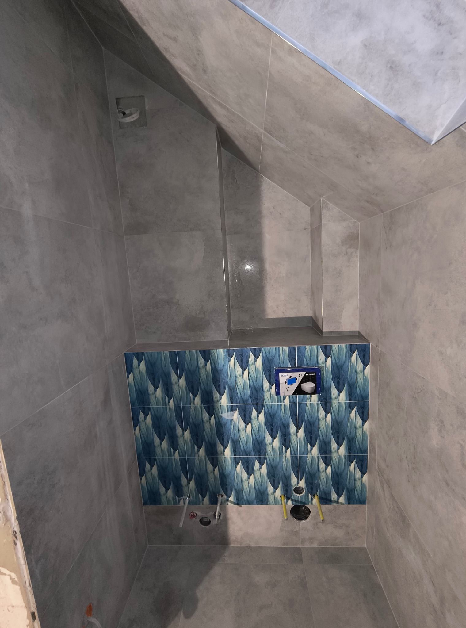 Ściana łazienki wyłożona szarymi płytkami imitującymi beton, z fragmentem dekoracyjnej ściany z niebieskimi płytkami w motywy piór, widoczne otwory na instalacje sanitarne i zamontowana podtynkowa...