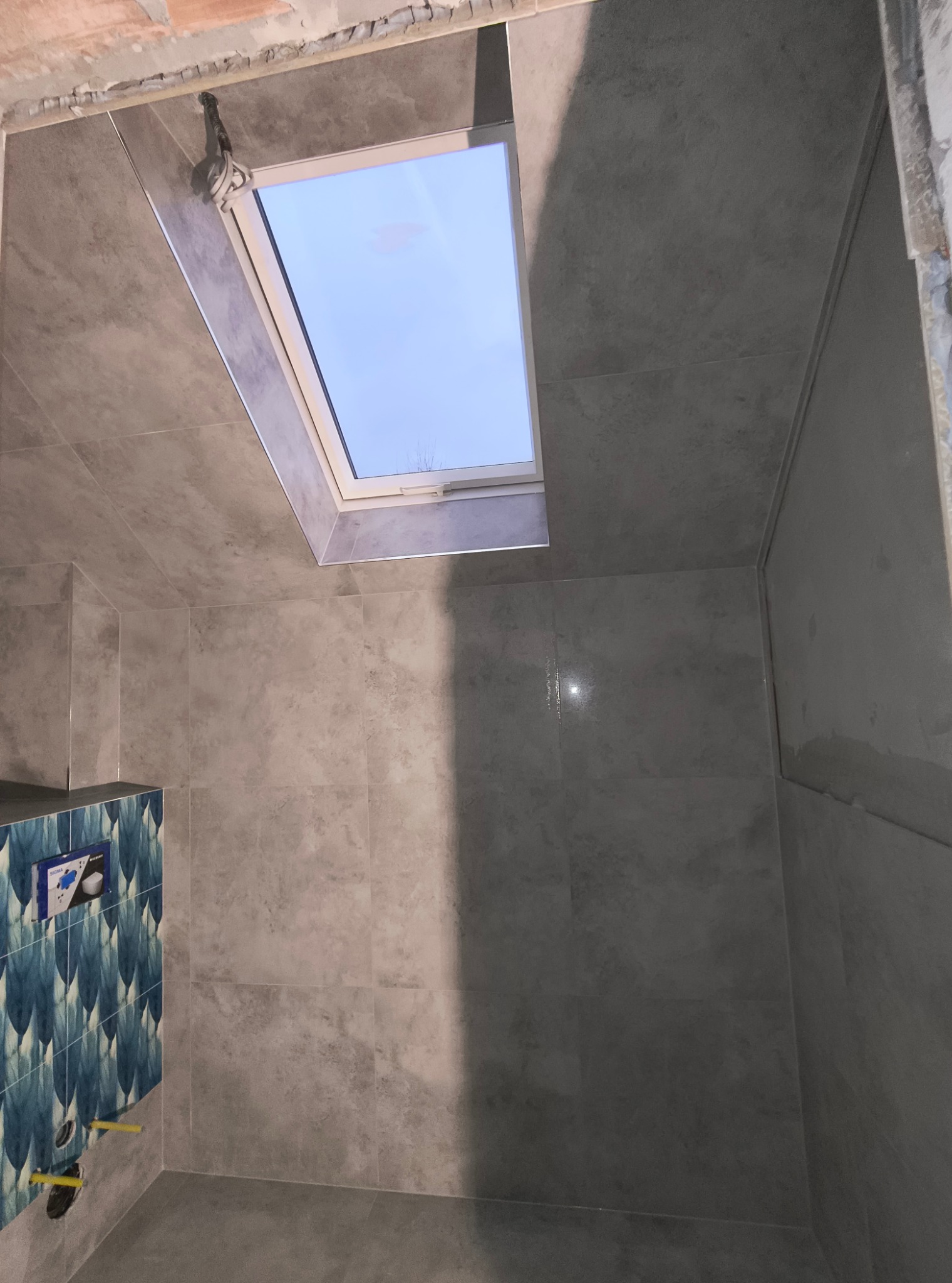 Szara łazienka w Limanowej z oknem dachowym i częściowo wykończoną ścianą z niebieskimi wzorzystymi płytkami, widoczne instalacje podtynkowe.
