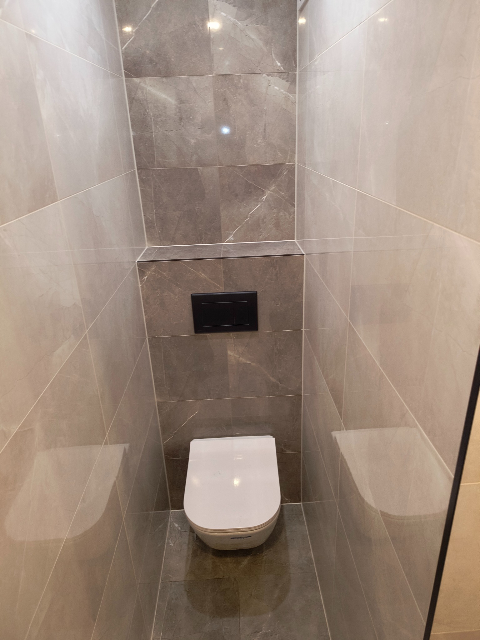 Wąska toaleta w nowoczesnej łazience wykończonej płytkami imitującymi marmur w odcieniach szarości, z podtynkową spłuczką z czarnym panelem.