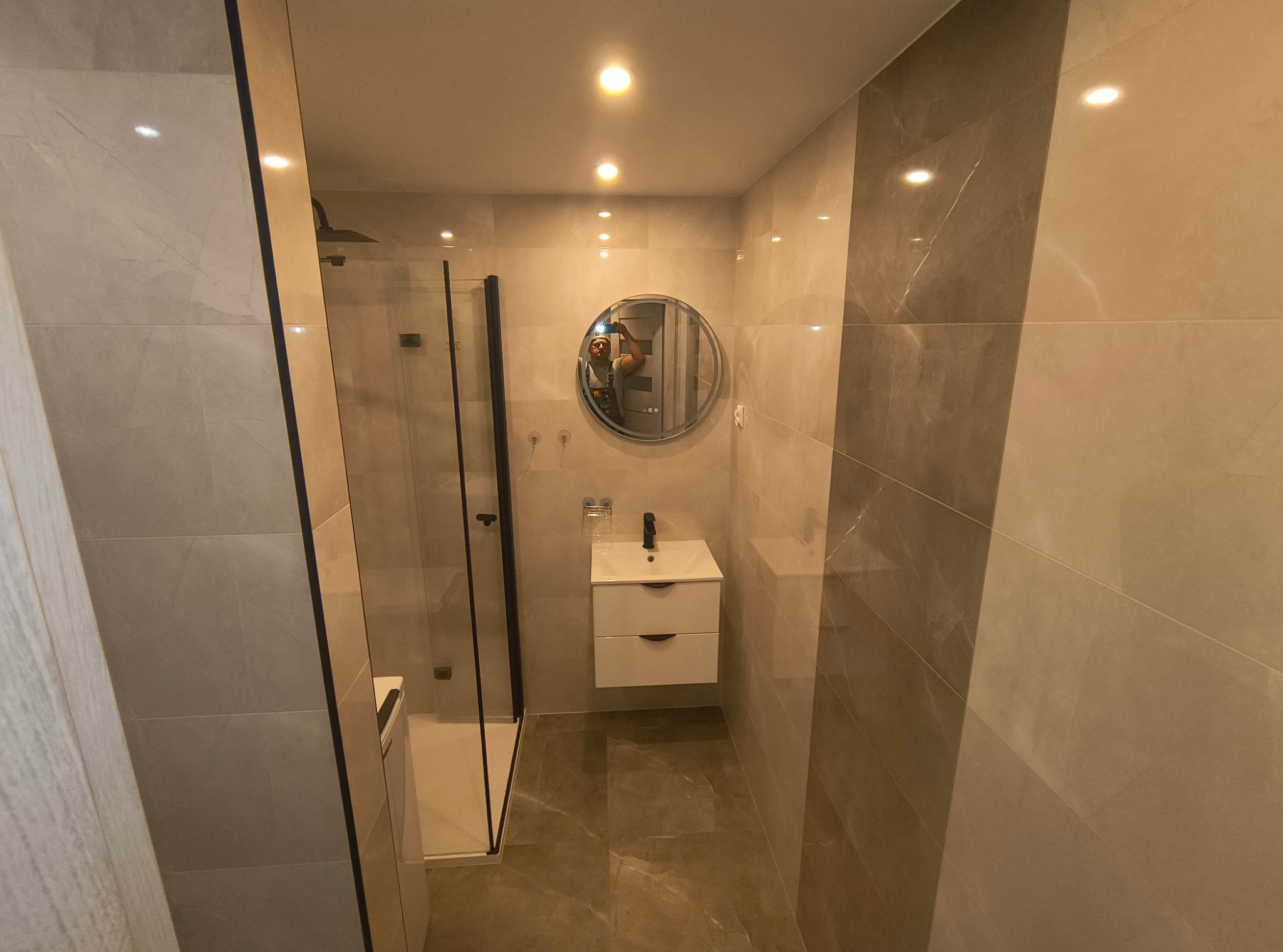 Nowoczesna łazienka z kabiną prysznicową typu walk-in, umywalką nablatową z szafką wiszącą i dużym okrągłym lustrem, wykończona płytkami w odcieniach beżu i szarości z widocznym odbiciem fotografa...