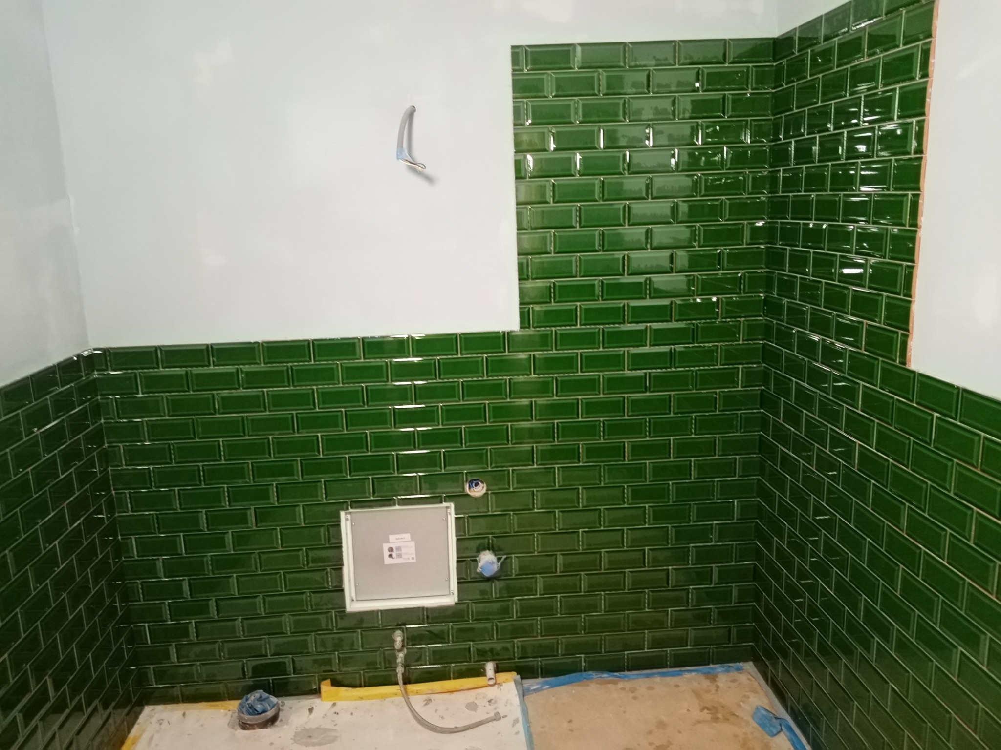 Ściany łazienki wyłożone błyszczącymi, zielonymi płytkami typu metro, częściowo zakończone, widoczne otwory na instalacje, surowa podłoga przed fugowaniem.