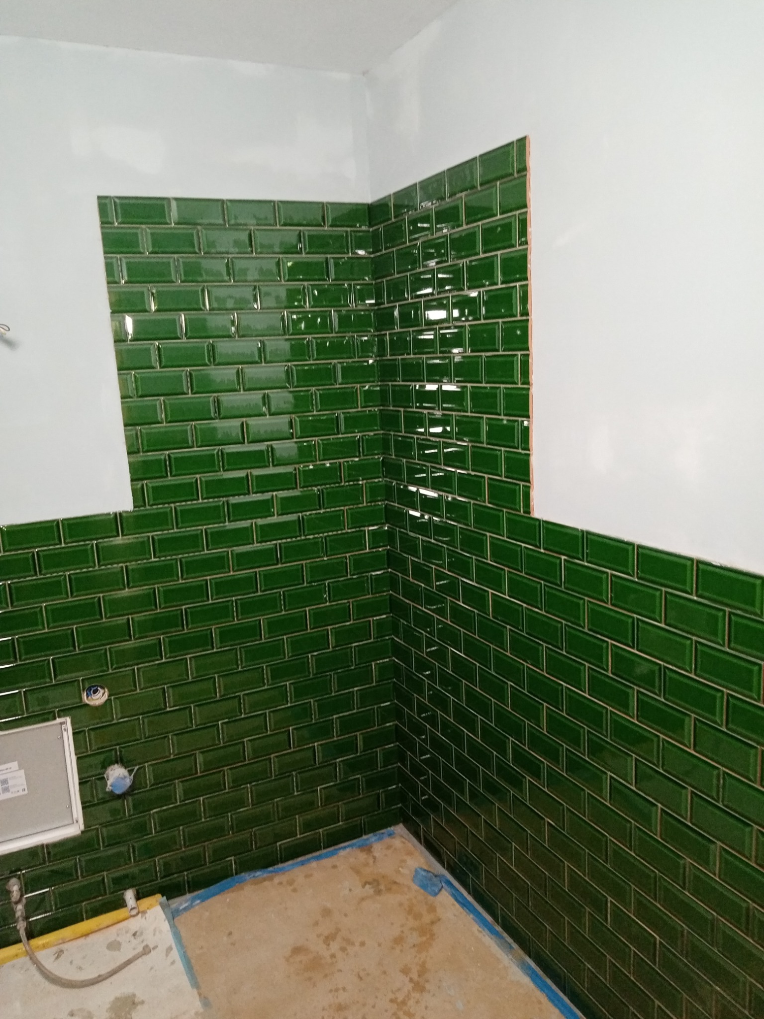 Ściana w trakcie układania ciemnozielonych, błyszczących płytek typu metro, z widocznymi fragmentami białej ściany i przygotowanym podłożem podłogowym.