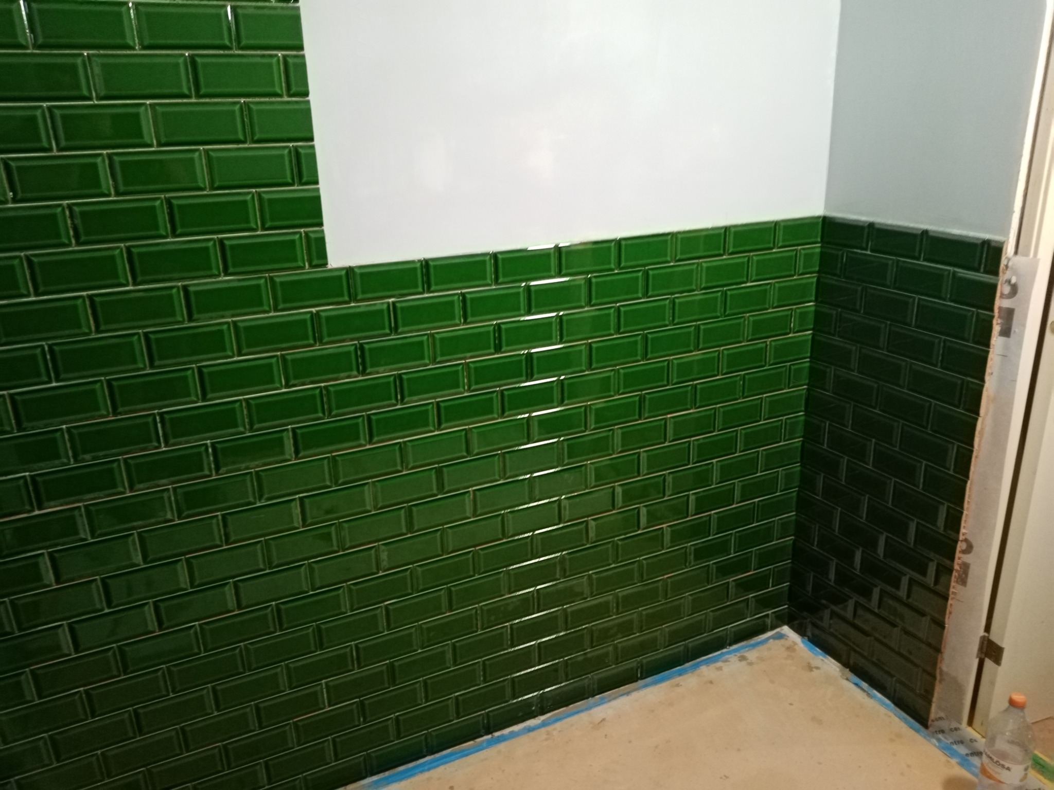 Ściana wyłożona błyszczącymi, ciemnozielonymi płytkami typu metro, częściowo pomalowana na biało, z widocznym fragmentem podłogi zabezpieczonej niebieską taśmą malarską.
