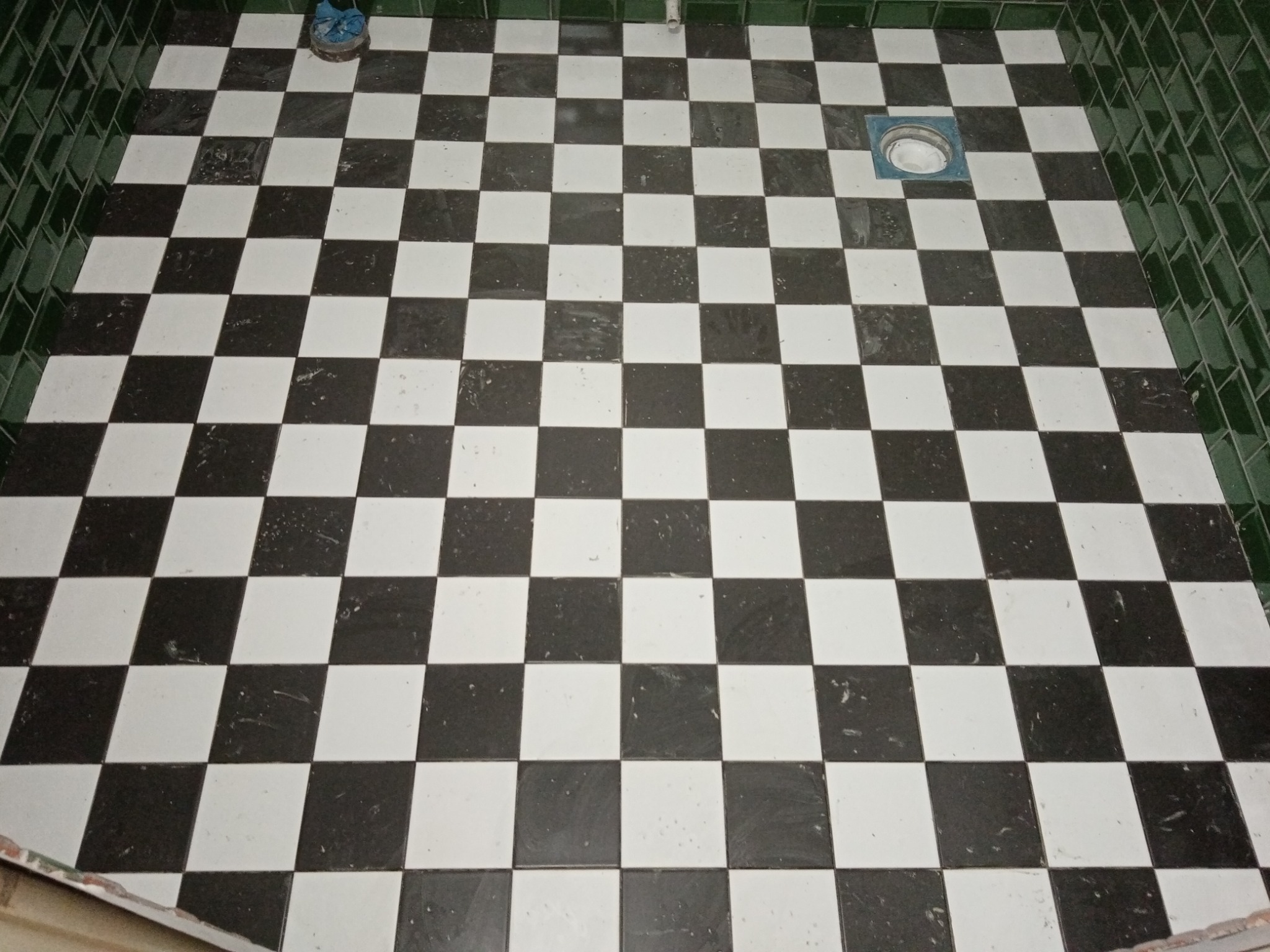 Podłoga w szachownicę z czarno-białych płytek ceramicznych, ciemnozielone płytki na ścianie, widoczny odpływ podłogowy i niebieskie buty robocze w rogu.