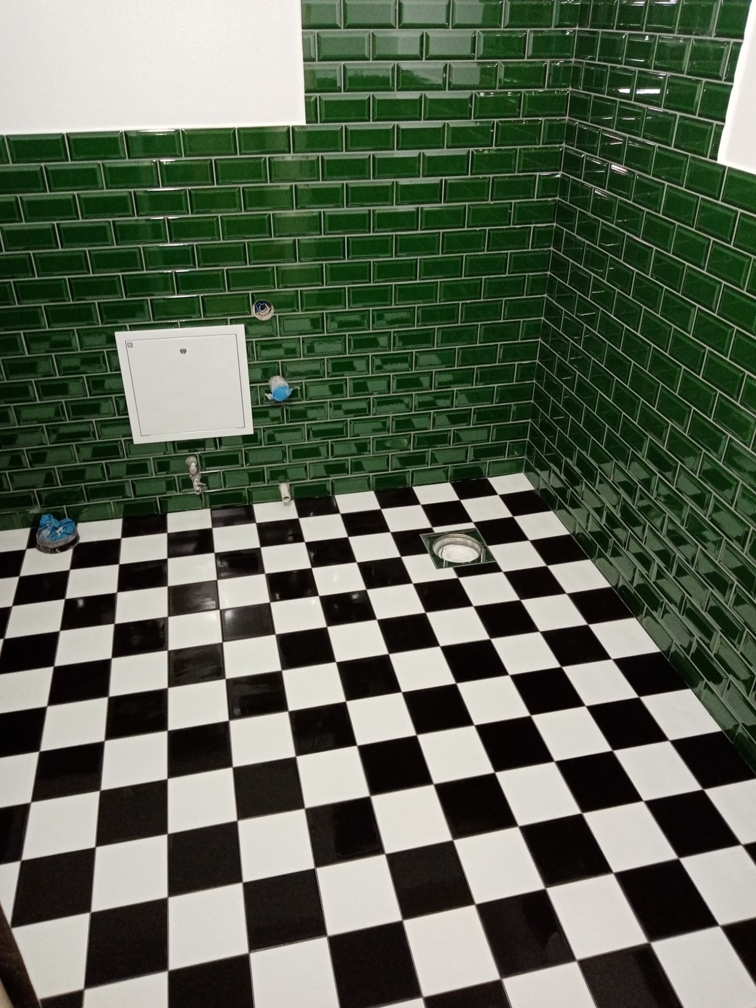 Świeżo wykafelkowana łazienka z błyszczącymi, szmaragdowozielonymi płytkami typu metro na ścianach i podłogą w czarno-białą szachownicę, widoczne przyłącza wodne i rewizja.