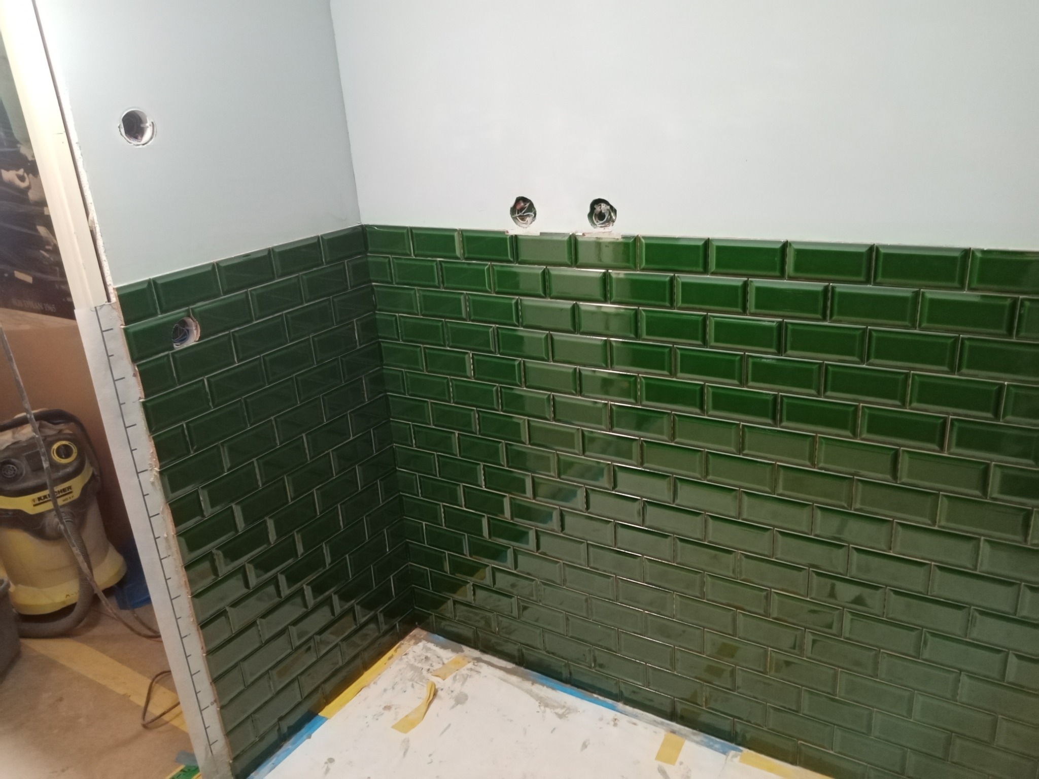 Ściana w trakcie układania ciemnozielonych, błyszczących płytek typu metro, widoczne otwory na instalacje elektryczne, na podłodze resztki zaprawy, w tle odkurzacz przemysłowy.