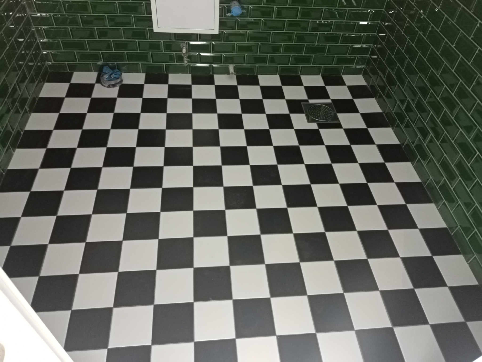 Ukończona podłoga w łazience z czarno-białą szachownicą, zielone, błyszczące płytki na ścianach i odpływ liniowy w rogu.