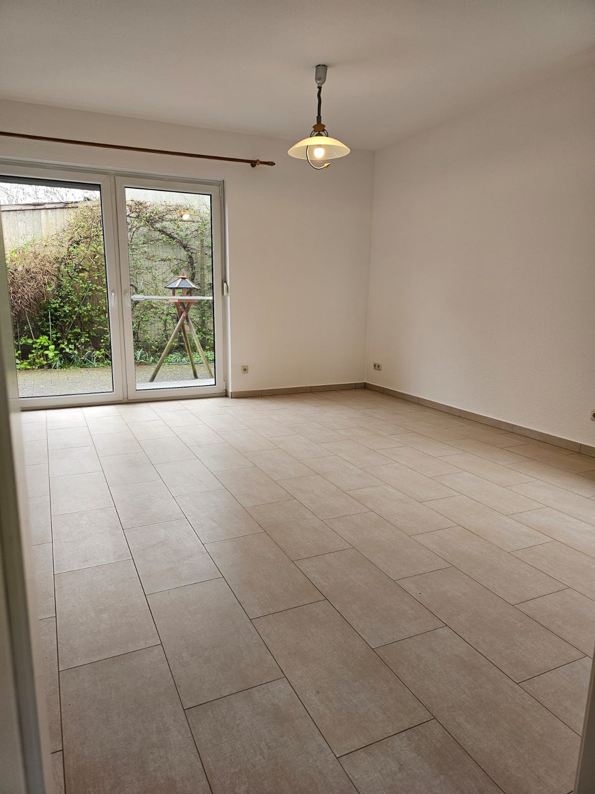 Puste pomieszczenie z beżowymi płytkami podłogowymi, widok na ogród przez przeszklone drzwi balkonowe, surowa żarówka wisząca na suficie.
