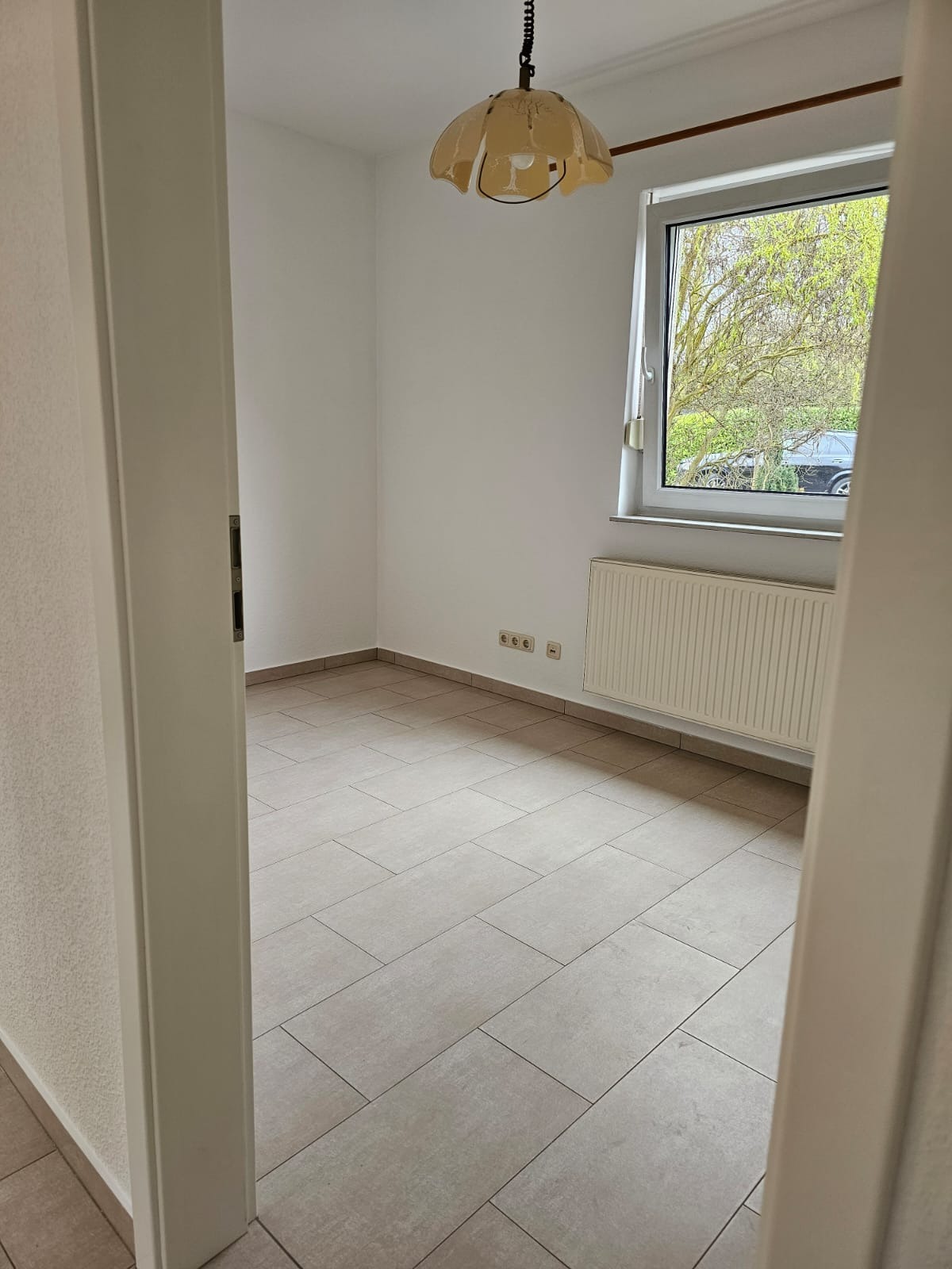 Pusty pokój z beżowymi płytkami podłogowymi, białymi ścianami, grzejnikiem pod oknem i lampą w stylu retro