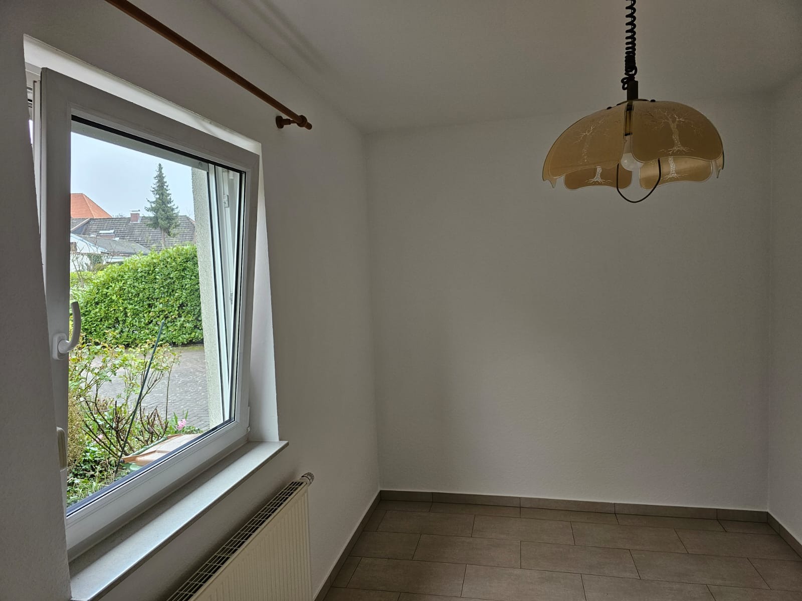 Pusty pokój z oknem wychodzącym na zieleń, wiszącą lampą w stylu retro i grzejnikiem pod oknem, przygotowany do malowania.