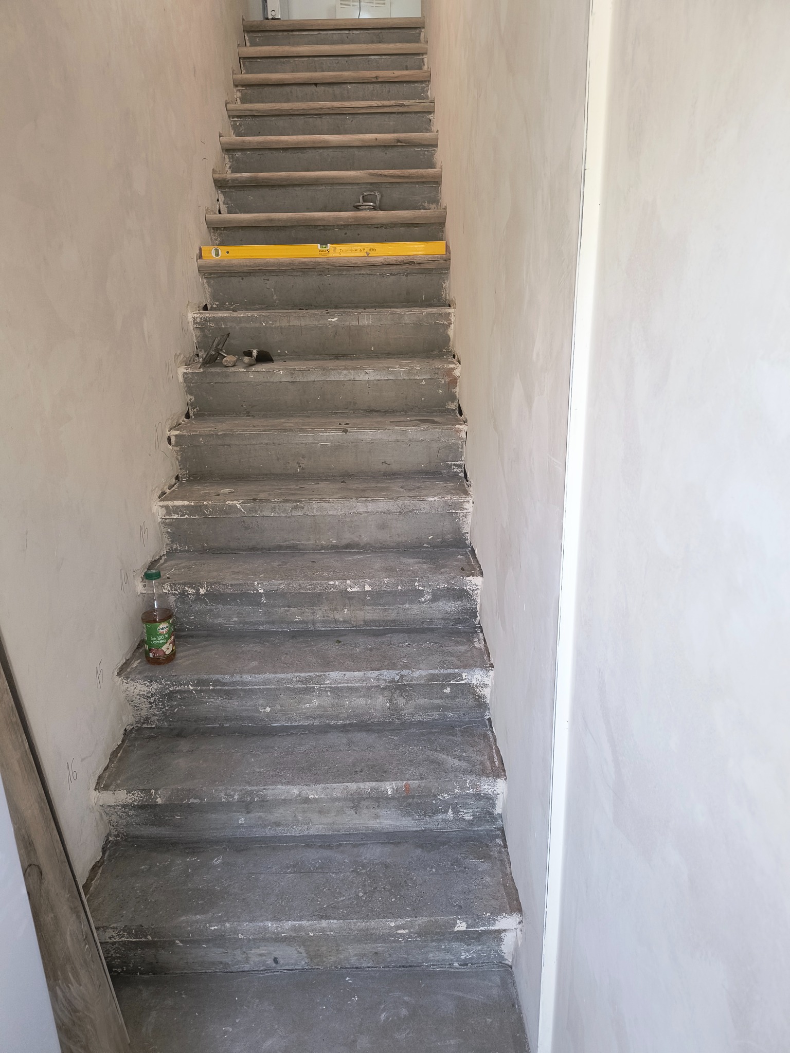 Surowe, betonowe schody w trakcie remontu, z poziomnicą na jednym ze stopni i butelką soku jabłkowego u podstawy, ściany pokryte świeżą gładzią.