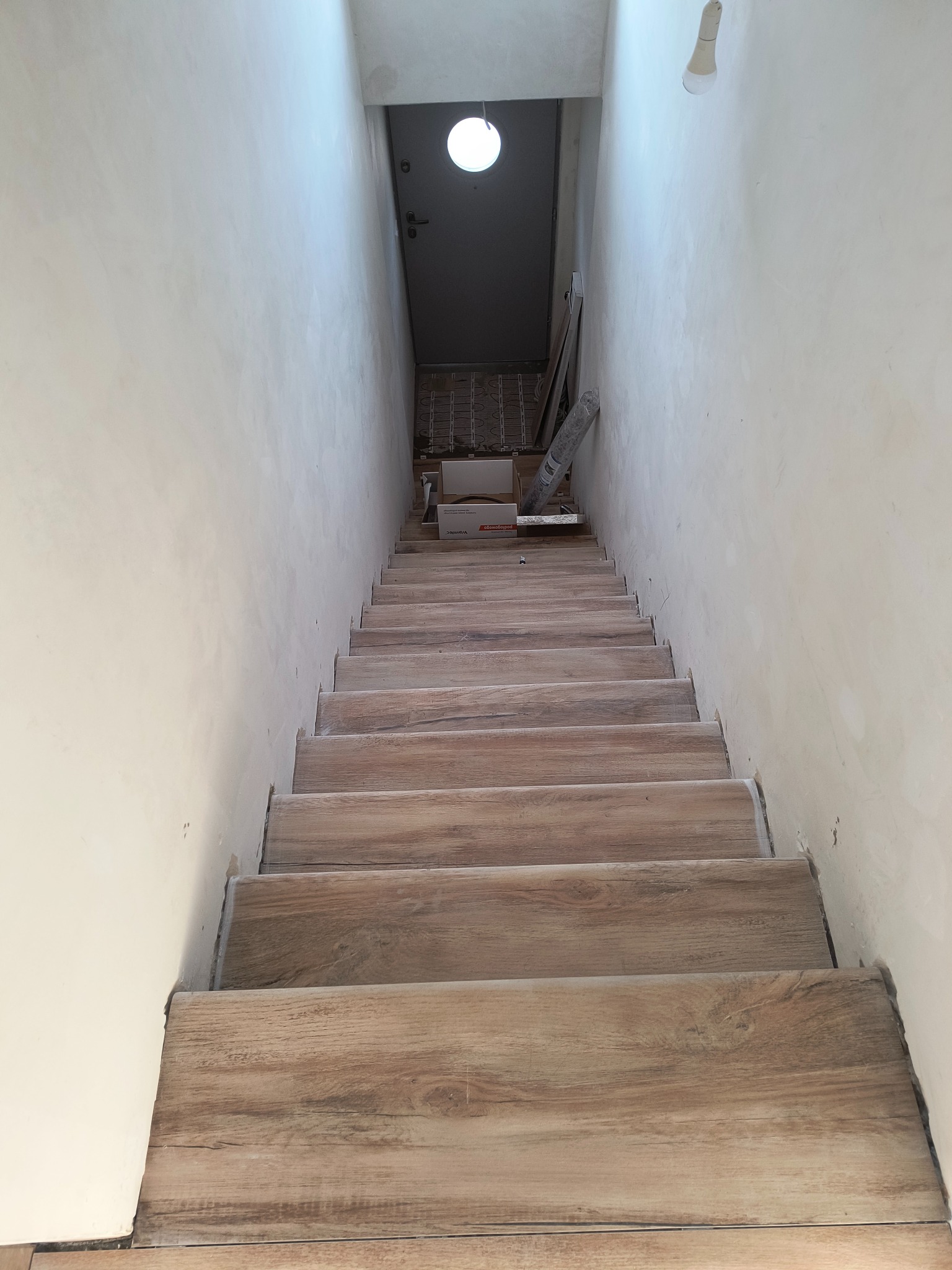 Schody wyłożone płytkami imitującymi drewno, prowadzące w dół, ściany przygotowane do malowania, widoczna surowa żarówka i materiały budowlane na końcu schodów.