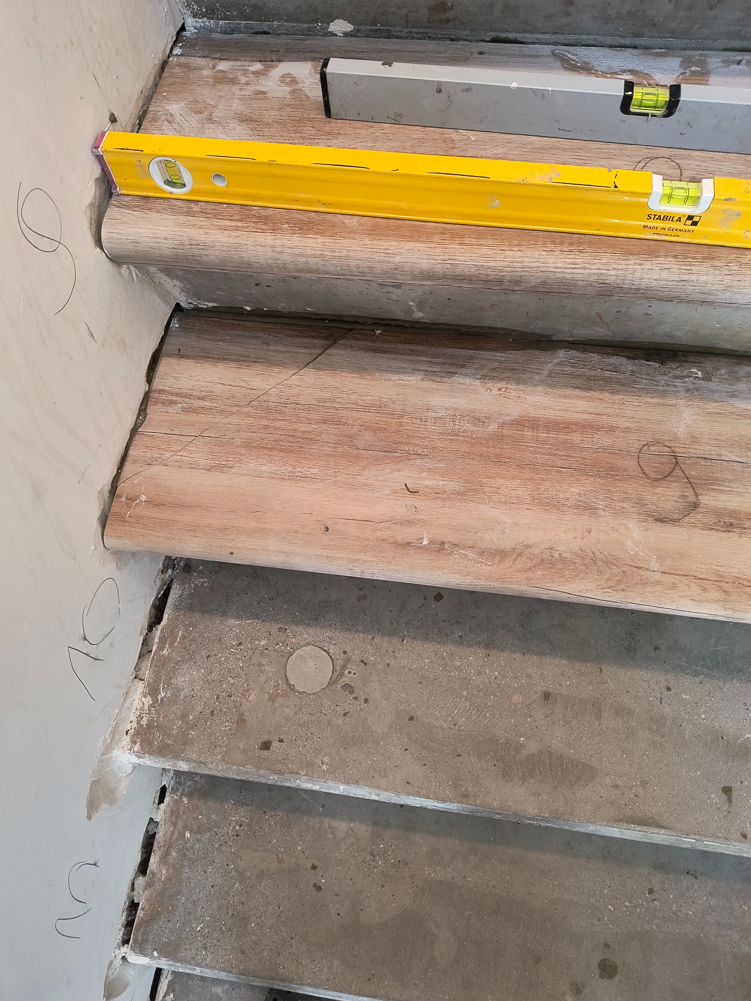 Montaż drewnopodobnych paneli na schodach betonowych, poziomowane żółtą poziomicą Stabila, widoczne oznaczenia ołówkiem na ścianie.