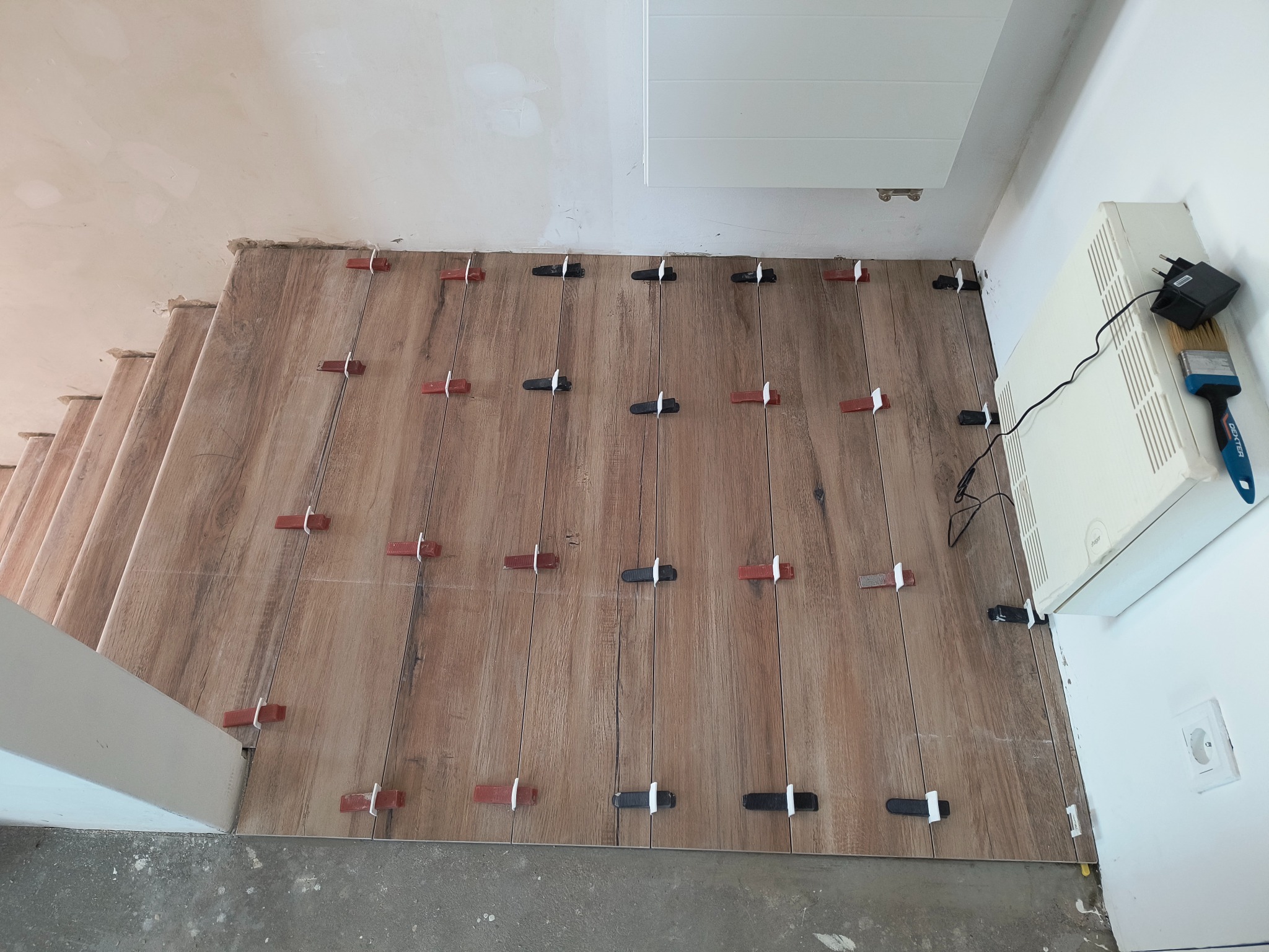 Układanie płytek imitujących drewno na schodach, z użyciem klipsów poziomujących w kolorach czerwonym i czarnym, widok z góry, fragment grzejnika na ścianie.