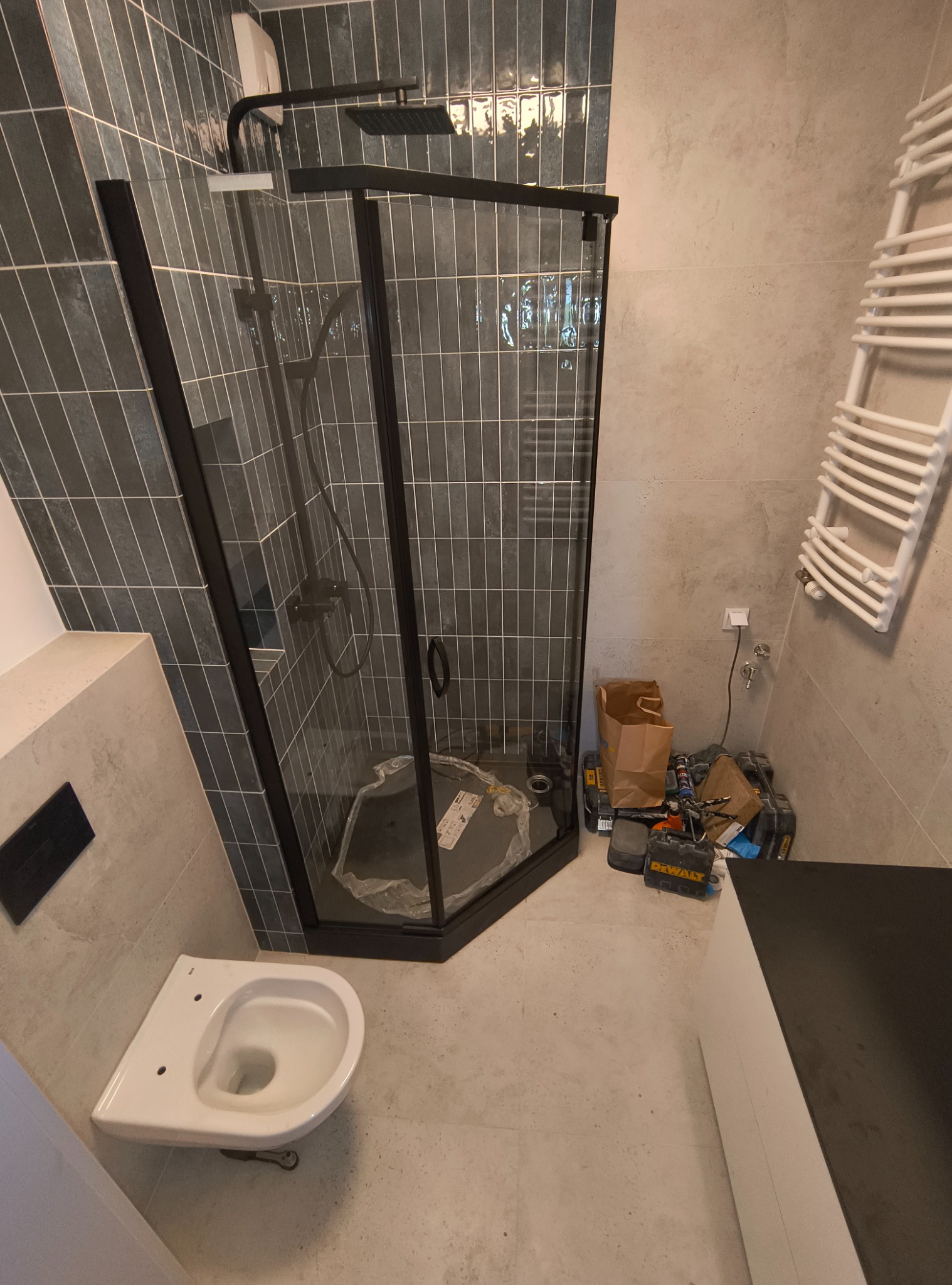 Nowoczesna kabina prysznicowa z czarnymi profilami i deszczownicą, częściowo zamontowana toaleta, grzejnik drabinkowy i narzędzia w trakcie prac wykończeniowych w łazience.