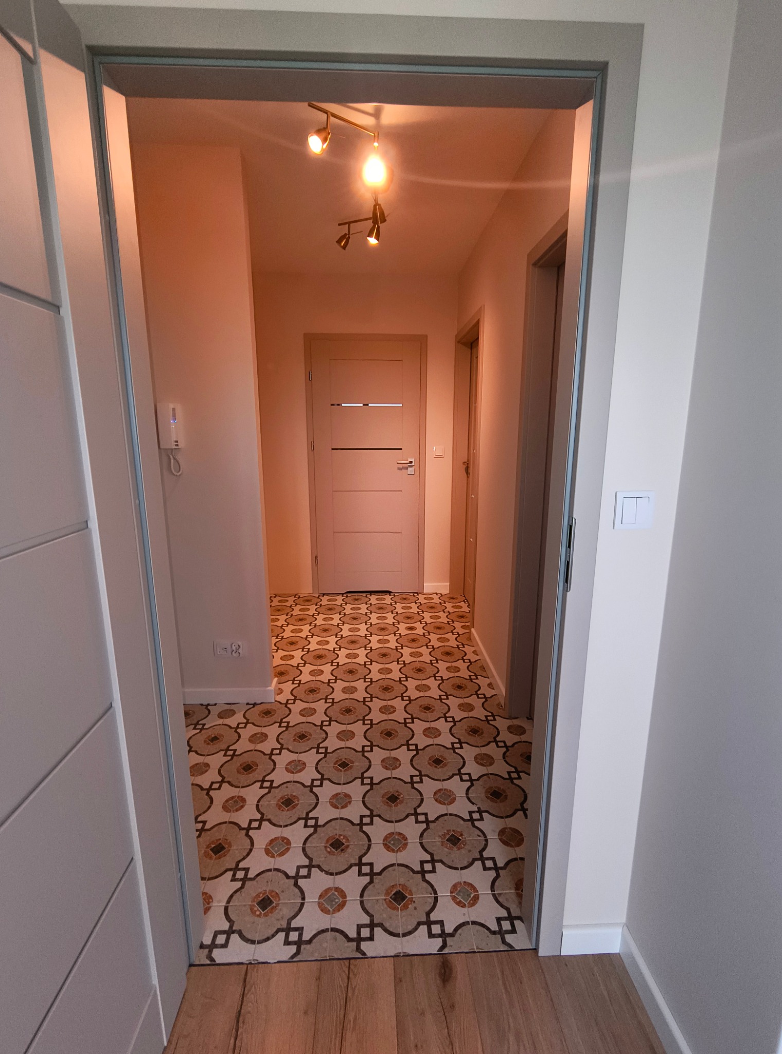 Wnętrze korytarza z wzorzystą podłogą, widoczne przez otwarte drzwi. Jasne ściany, oświetlenie sufitowe i fragment drewnianej podłogi przy wejściu.