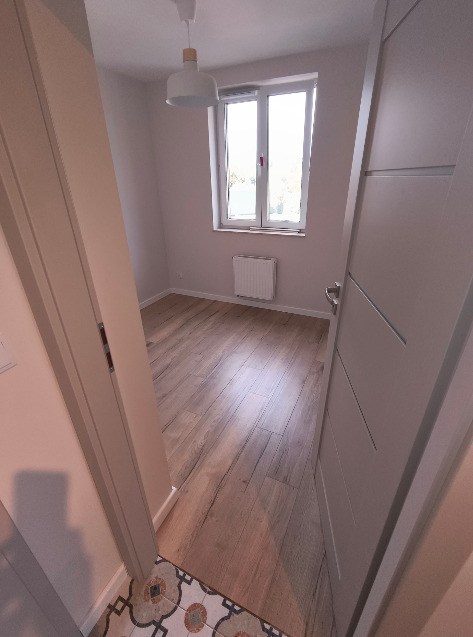 Jasne wnętrze pokoju z drewnianą podłogą, widokiem na okno i grzejnik. Drzwi wewnętrzne w kolorze szarym są częściowo otwarte. Dekoracyjne płytki podłogowe przy wejściu.