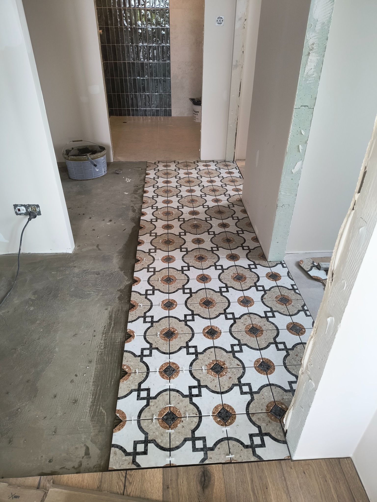 Wnętrze w trakcie prac wykończeniowych: ułożona częściowo wzorzysta terakota na podłodze, w tle ciemne płytki na ścianie. Widoczne narzędzia i materiały budowlane.