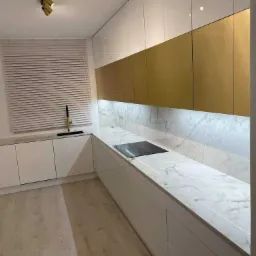 Nowoczesna kuchnia z białymi szafkami, złotymi górnymi szafkami i marmurowym blatem z wbudowaną płytą grzewczą, oświetlona listwą LED pod szafkami.