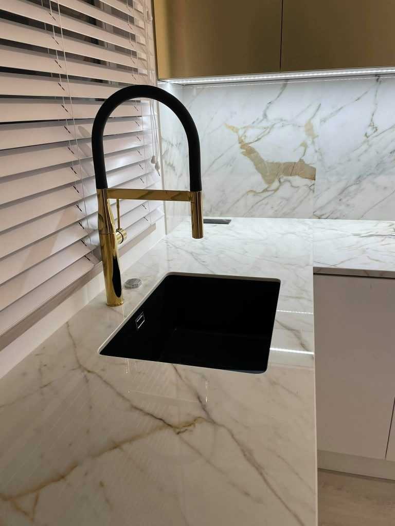 Czarny zlew wpuszczony w jasny blat z marmurowym wzorem, bateria w kolorze złota z czarnym uchwytem, żaluzje w tle.