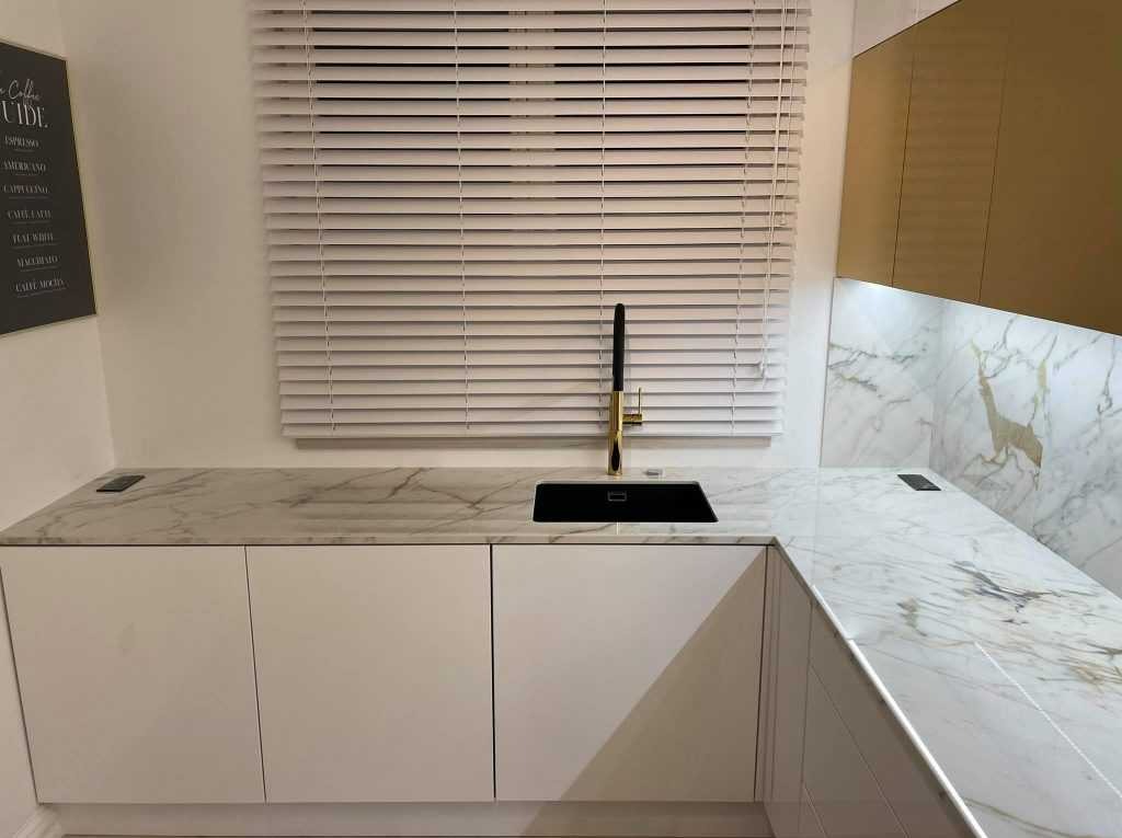 Kuchenny blat z marmuru z czarnym zlewem i złotą baterią na tle białych szafek i żaluzji okiennych.