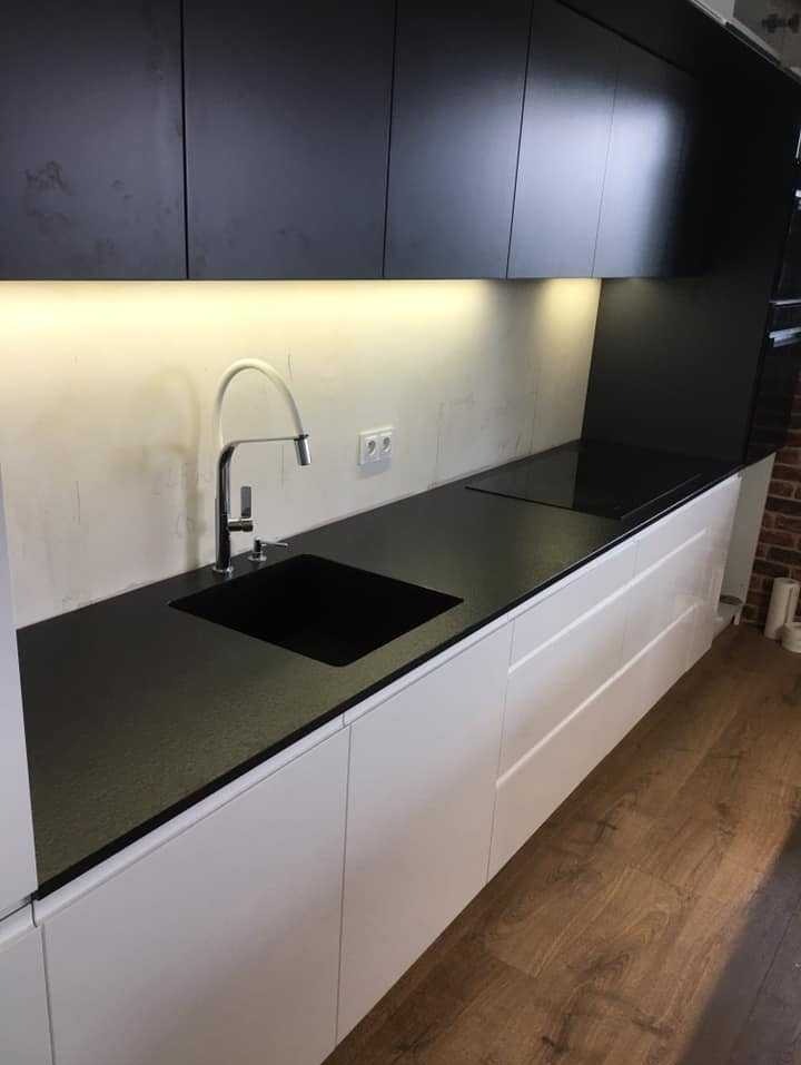 Nowoczesna kuchnia z czarnym blatem, białymi szafkami i chromowaną baterią, oświetlona listwą LED pod szafkami wiszącymi, widoczny fragment ściany z cegły.