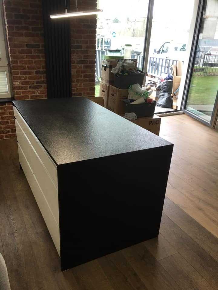 Czarny blat kamienny na białej szafce w pomieszczeniu z ceglaną ścianą i kartonami w tle, widoczne przesuwne drzwi balkonowe.