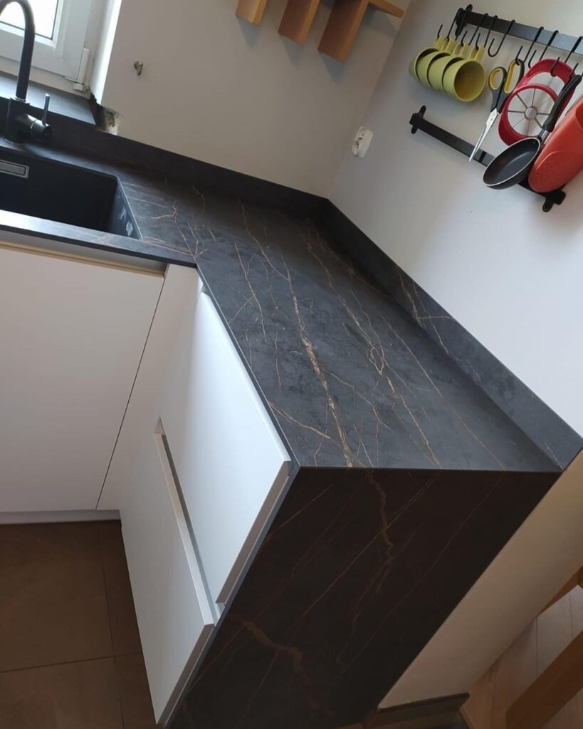Czarny blat kuchenny z marmurkowym wzorem w odcieniach złota, zamontowany na białych szafkach kuchennych, widoczny z perspektywy lekko z góry, z czarnym zlewem po lewej stronie i akcesoriami...