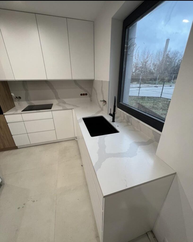 Nowoczesna kuchnia z białymi szafkami i blatem z wzorem imitującym marmur, czarny zlew i bateria, duże okno z widokiem na zewnątrz.