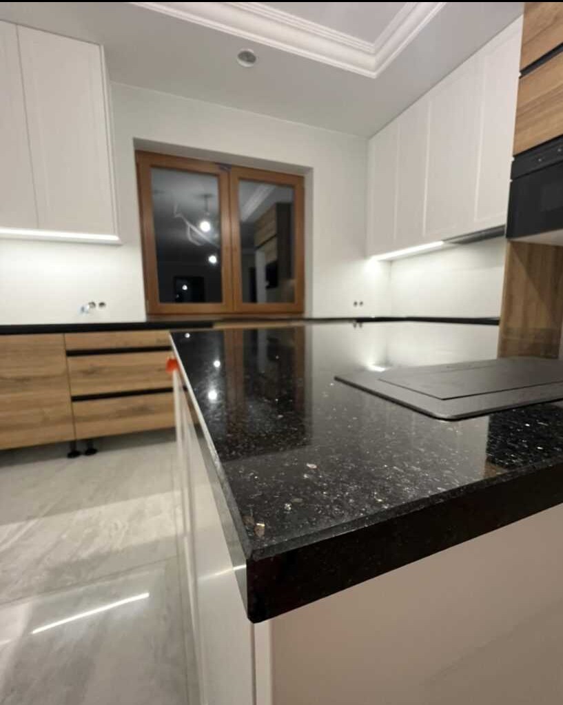 Błyszczący, czarny blat z drobnymi, jasnymi inkluzjami, wkomponowany w nowoczesną kuchnię z białymi szafkami i drewnianymi akcentami. Widoczna płyta grzewcza i odbicie światła w polerowanej...