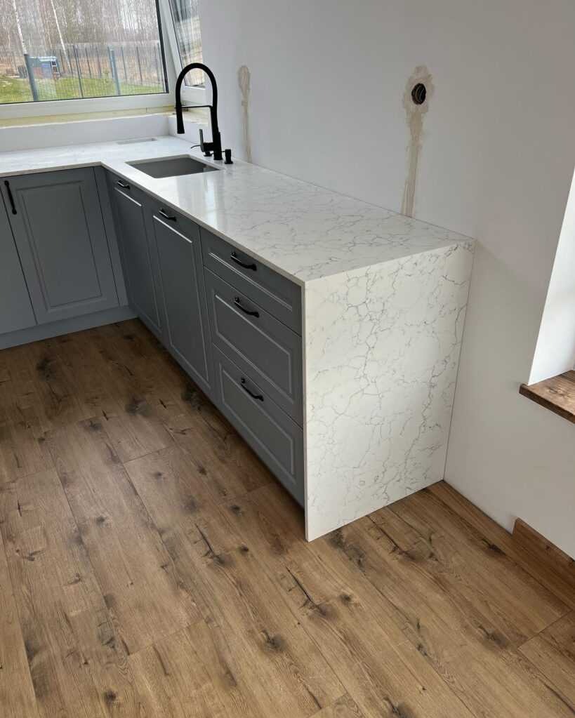 Szary zestaw szafek kuchennych z blatem z białego konglomeratu z szarymi żyłkami, czarna bateria kuchenna, drewniana podłoga, widoczne ślady po instalacji na ścianie.