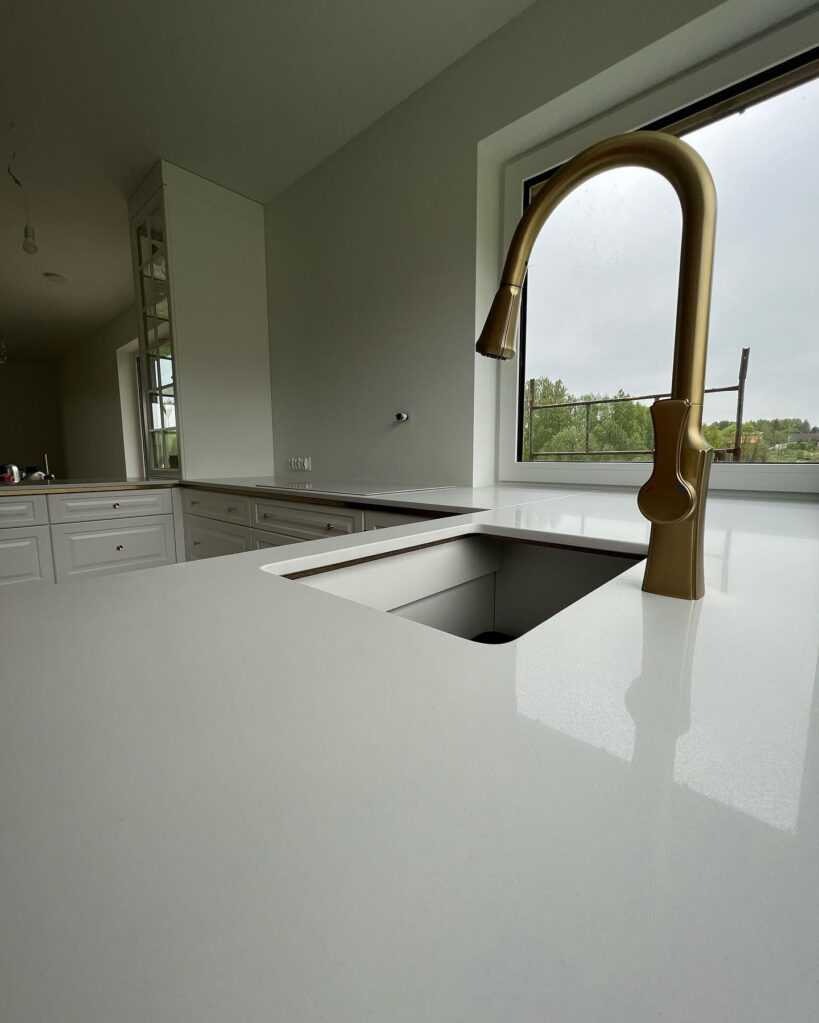 Jasny blat kuchenny z konglomeratu z wbudowanym zlewem i złotą baterią na tle okna z widokiem na zieleń.