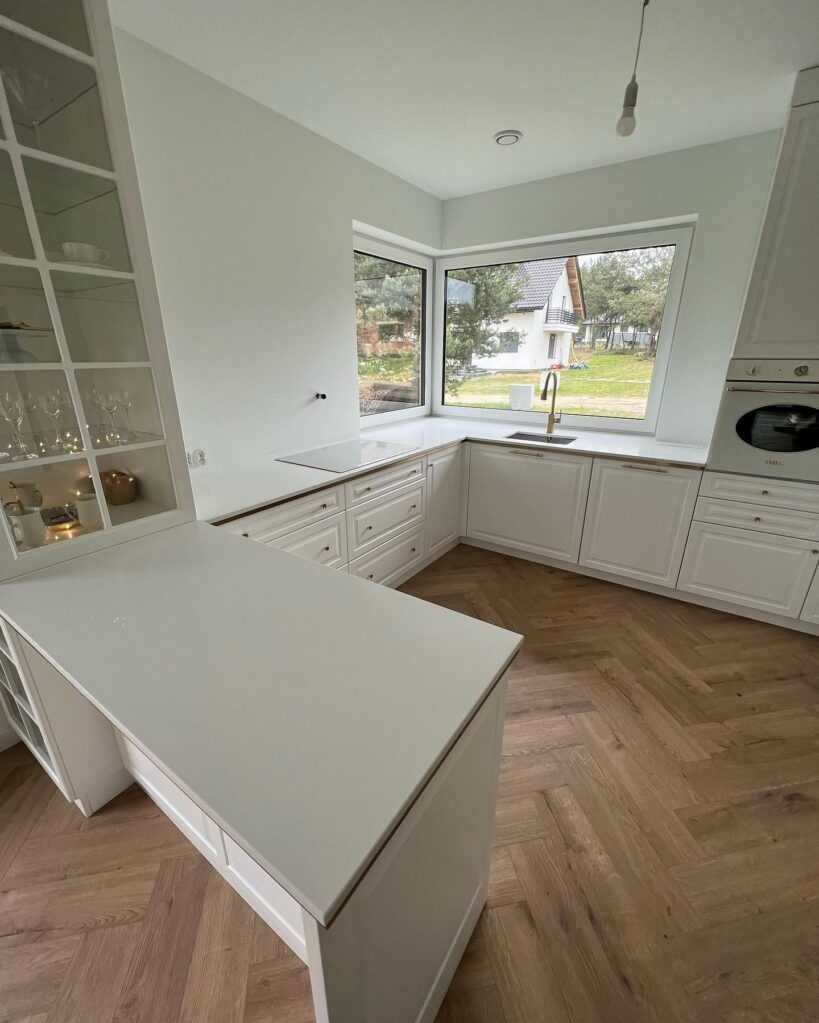 Jasna kuchnia z białymi szafkami w stylu klasycznym, drewnianą podłogą ułożoną w jodełkę i wyspą z blatem o jasnym odcieniu, widok na ogród przez duże okno.