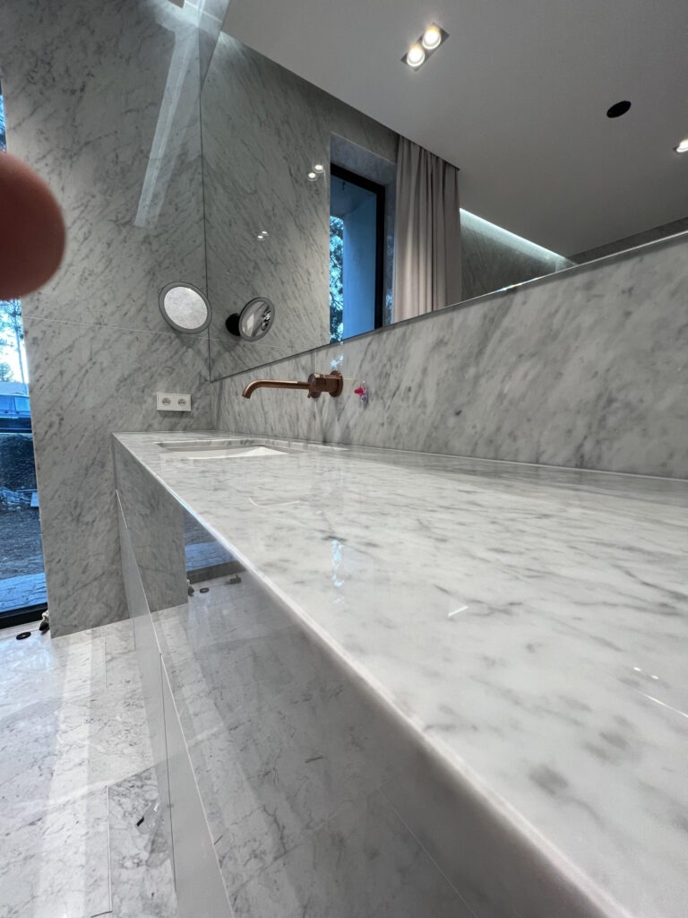 Biały marmurowy blat łazienkowy z miedzianą armaturą, podświetlane lustro i marmurowe ściany w nowoczesnej łazience.