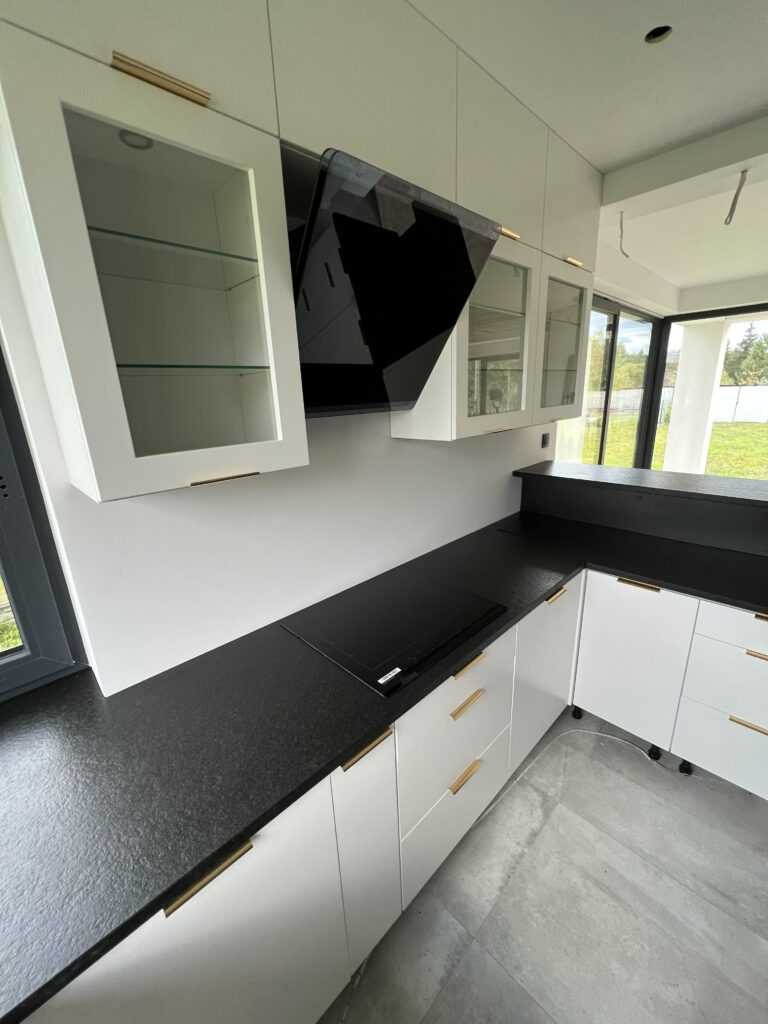 Czarny blat kamienny w nowoczesnej kuchni w Tarnobrzegu, z białymi szafkami i złotymi uchwytami, widok na płytę indukcyjną i okno.