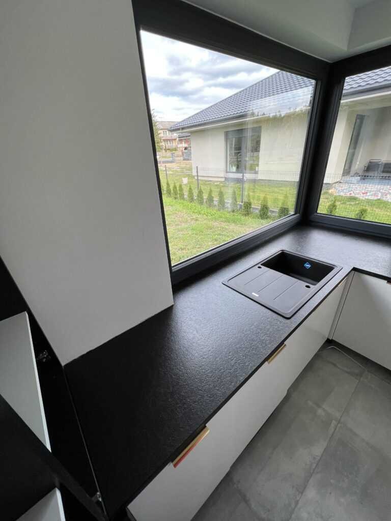 Czarny, teksturowany blat kuchenny z wbudowanym zlewozmywakiem, zamontowany przy oknie z widokiem na ogród i dom.