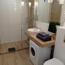 Nowoczesna łazienka z kabiną prysznicową typu walk-in, umywalką nablatową na drewnianym blacie nad pralką, toaletą i dekoracyjnym oświetleniem w postaci żyrandola.