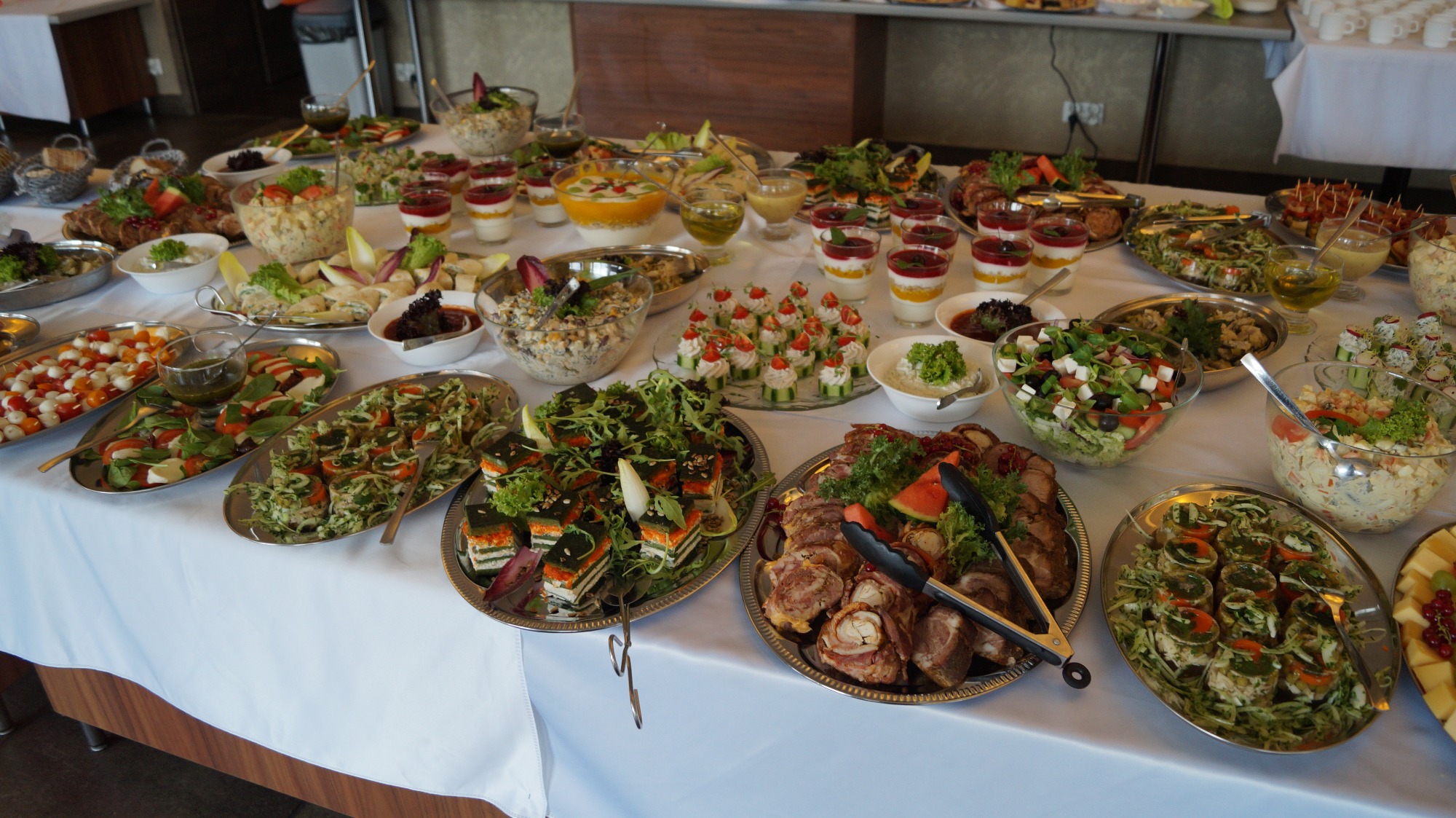 Bogato zastawiony stół cateringowy z różnorodnymi przystawkami: sałatki, roladki mięsne z szczypcami, mini kanapeczki, desery w szklankach i inne przekąski na srebrnych paterach.