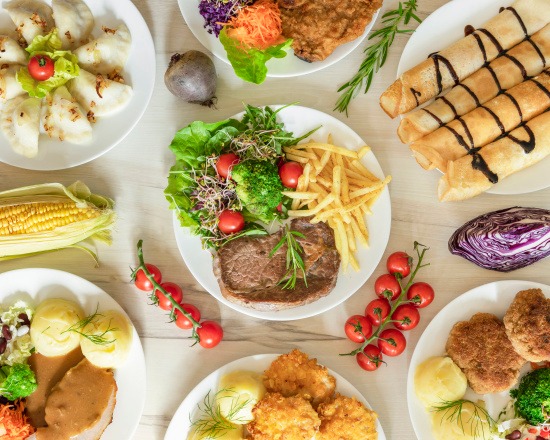 Widok z góry na stół zastawiony różnorodnymi daniami: pierogi, kotlet, naleśniki z polewą czekoladową, stek z frytkami i sałatką, pieczeń z sosem i ziemniakami, kotlety mielone z ziemniakami...