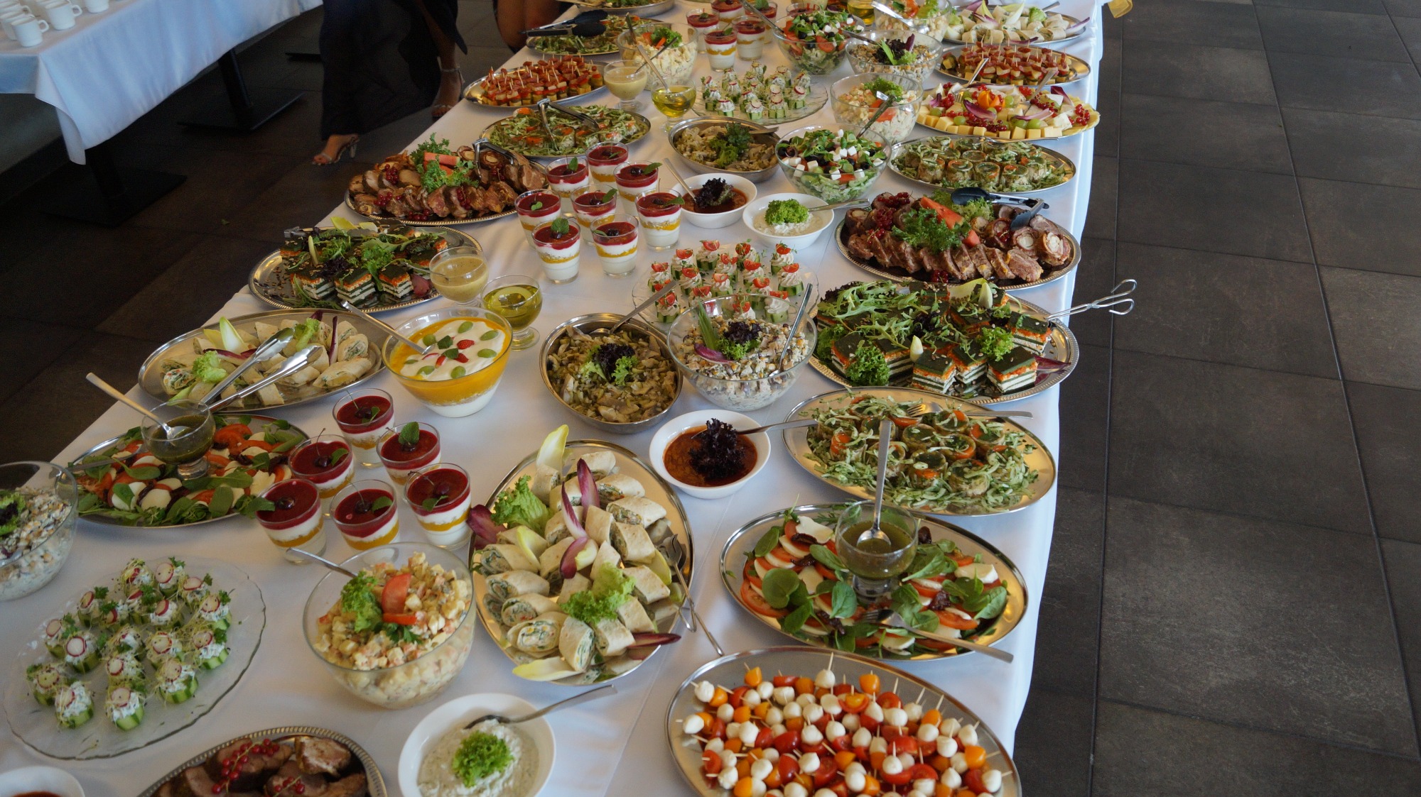 Bogato zastawiony stół z różnorodnymi przekąskami i daniami, w tym sałatkami, roladkami, deserami w szklankach, koreczkami caprese i mięsami, prezentowanymi na srebrnych paterach i w szklanych...