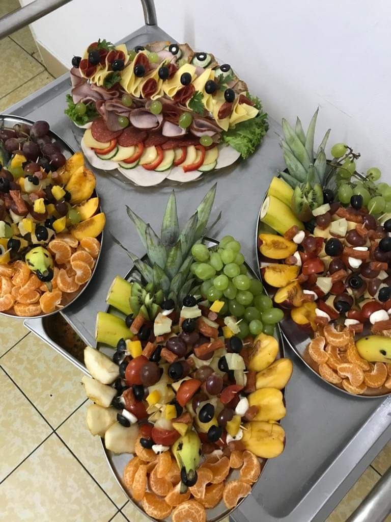 Cztery srebrne tace z kompozycjami z owoców, wędlin i serów, ułożone na wózku cateringowym na tle kafelkowej podłogi.