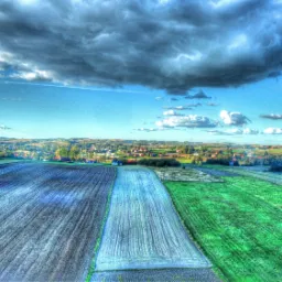 Szeroki kadr z drona na pola uprawne, w tle zabudowania wiejskie pod błękitnym niebem z ciemnymi chmurami, wykonany przez dronteamrzeszow.pl.