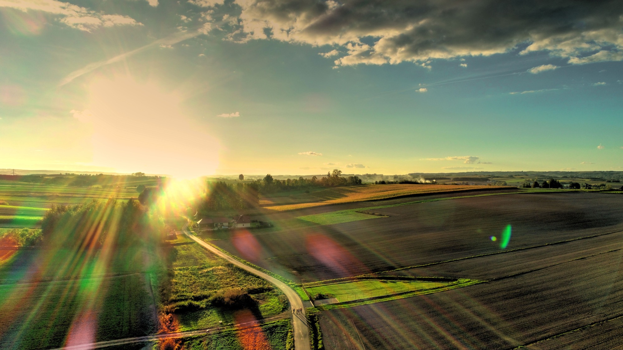 Panoramiczny widok z drona na polskie pola uprawne o zachodzie słońca, z drogą i grupą spacerujących osób w oddali.