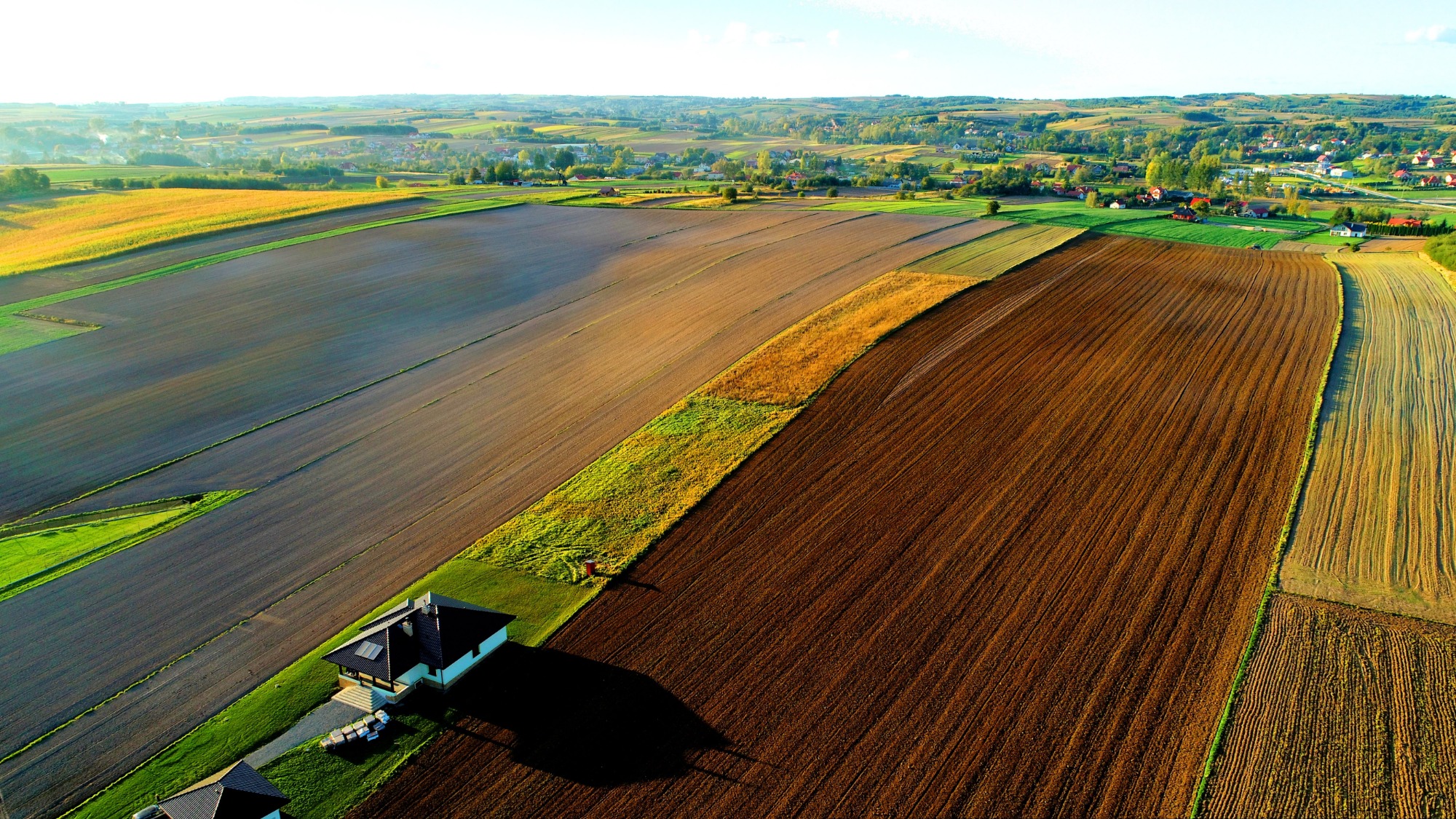 Panoramiczny widok z drona na wiejski krajobraz z polami uprawnymi o różnych kolorach i fakturach, domami w tle oraz cieniem rzucanym przez budynek w lewym dolnym rogu.