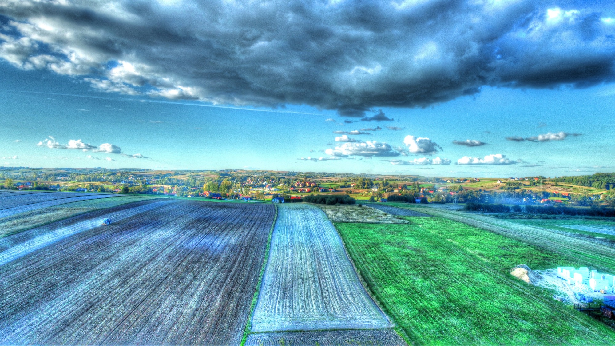 Szeroki kadr z drona na pola uprawne, w tle zabudowania wiejskie pod błękitnym niebem z ciemnymi chmurami, wykonany przez dronteamrzeszow.pl.
