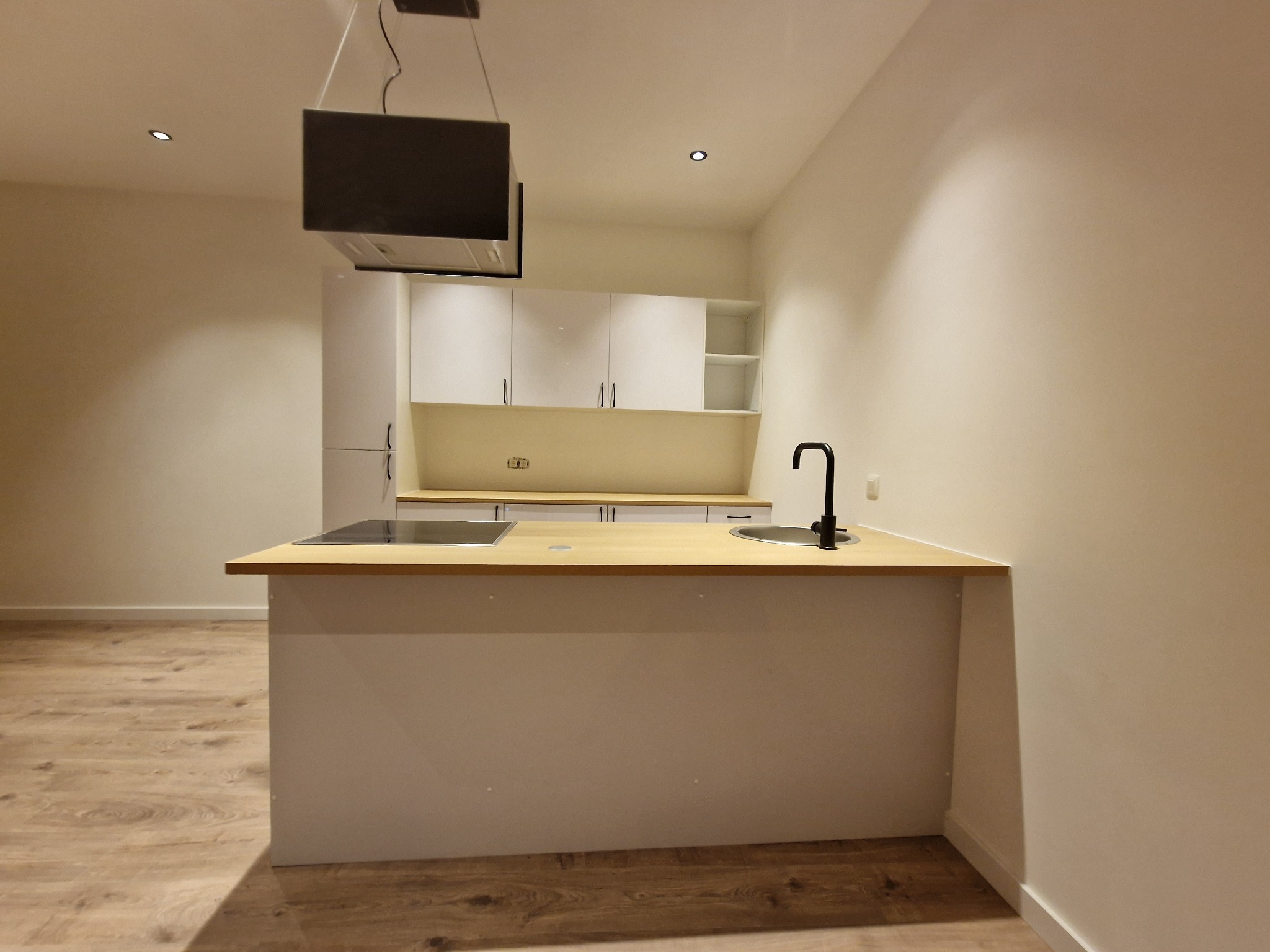 Nowoczesna, minimalistyczna kuchnia z wyspą, jasnymi szafkami i drewnianym blatem, czarna bateria kuchenna, oświetlenie punktowe, parkiet na podłodze.