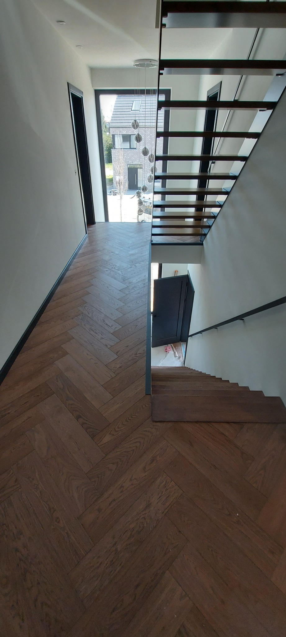 Wnętrze domu z parkietem ułożonym w jodełkę, schodami z drewnianymi stopniami i metalową balustradą oraz nowoczesnym oświetleniem wiszącym.