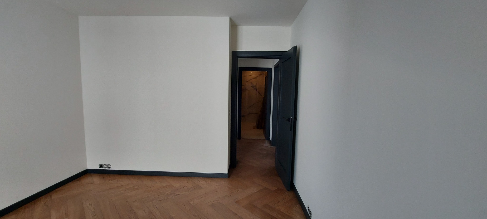Pusty pokój po odświeżeniu: białe ściany, ciemne listwy przypodłogowe i ościeżnice, parkiet w jodełkę. W tle widoczna łazienka z marmurowymi płytami.