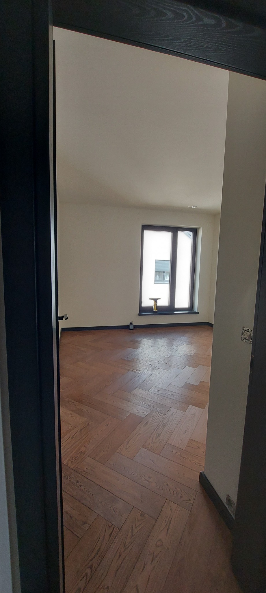 Pusty pokój z parkietem ułożonym w jodełkę, widok zza czarnej futryny drzwi, okno z dekoracyjnym elementem na parapecie.