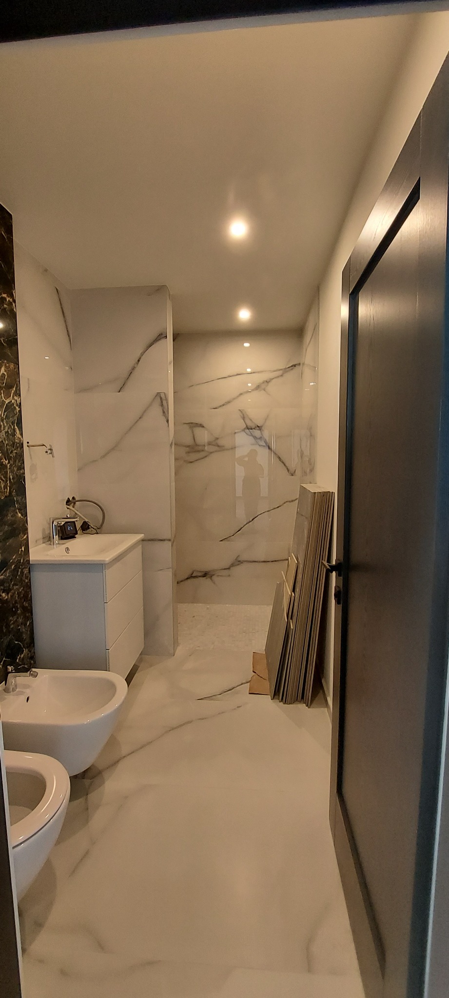 Nowoczesna łazienka w trakcie wykańczania z białymi płytkami imitującymi marmur, widoczne białe sanitariaty, umywalka z szafką oraz stos płytek opartych o ścianę przy otwartych drzwiach.